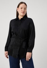 Black, long-sleeve shirt made of smooth fabric with a collared neckline, front button closure, and tied belt at the waist, paired with denim.