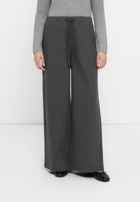 Hellebeinsthosen aus schattiertem grauem Stoff mit einem Zugbandbund. Glatte Textur mit seitlichen Schlitzen, kombiniert mit schwarzen flachen Schuhen.