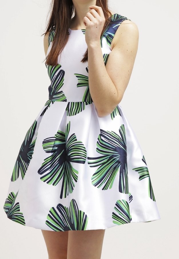 Femme portant une robe blanche sans manches avec un grand imprimé de feuilles tropicales vertes et noires, debout devant un fond uni.