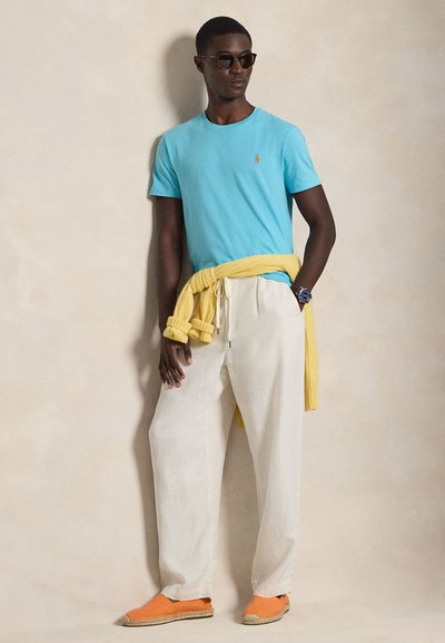 Ανοιχτό μπλε μπλουζάκι με μικρό λογότυπο, εκρού παντελόνι, κίτρινη μπλούζα δεμένη γύρω από τη μέση και πορτοκαλί εσπαντρίγιες στα πόδια.