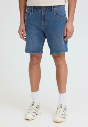 Homme portant un short en denim bleu, des chaussettes blanches montantes et des chaussures de sport blanches, debout devant un fond clair uni.