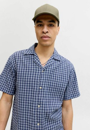 Junger Mann mit olivgrüner Mütze und blau kariertem Kurzarmhemd vor einfarbig hellem Hintergrund.