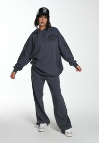 Sudadera de gran tamaño en gris oscuro con diseños florales, combinada con pantalones de chándal a juego y una gorra en blanco y negro. Presenta un bolsillo frontal.