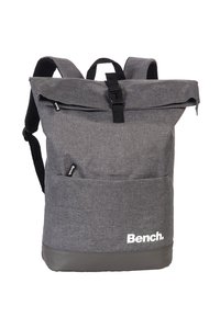 Sac à dos en tissu gris avec un design à rabat, présentant des accents noirs, des bretelles ajustables, des poches extérieures et un logo sur le devant.