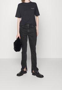 Maglietta nera in cotone con logo, abbinata a jeans neri aderenti con cerniere laterali e scarpe in pelle nera. Tiene una pochette nera in pelliccia.