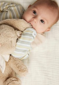 Bébé dans une tenue rayée, jaune clair et gris, tenant un jouet en peluche beige doux. L'arrière-plan présente un tissu texturé de couleur claire.