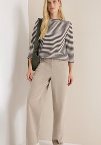 Femme portant un haut à rayures brun et blanc à manches longues, un pantalon large beige et portant un sac à épaule vert foncé, se tenant à l'intérieur.