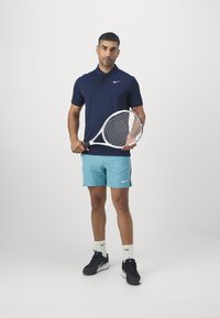 Polo de color marino y pantalones cortos de color azul claro con logotipos de Nike. Sostiene una raqueta de tenis, lleva zapatillas deportivas negras y calcetines blancos. Fondo minimalista.