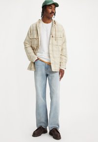 Beige geruite flanellen shirt, witte crewneck t-shirt eronder, lichtblauwe jeans, bruine veterschoenen en een groene pet.