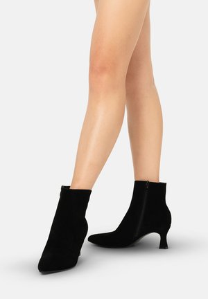 Piernas con botines de ante negro con cremalleras laterales y tacones bajos tipo kitten, sobre una superficie gris claro.