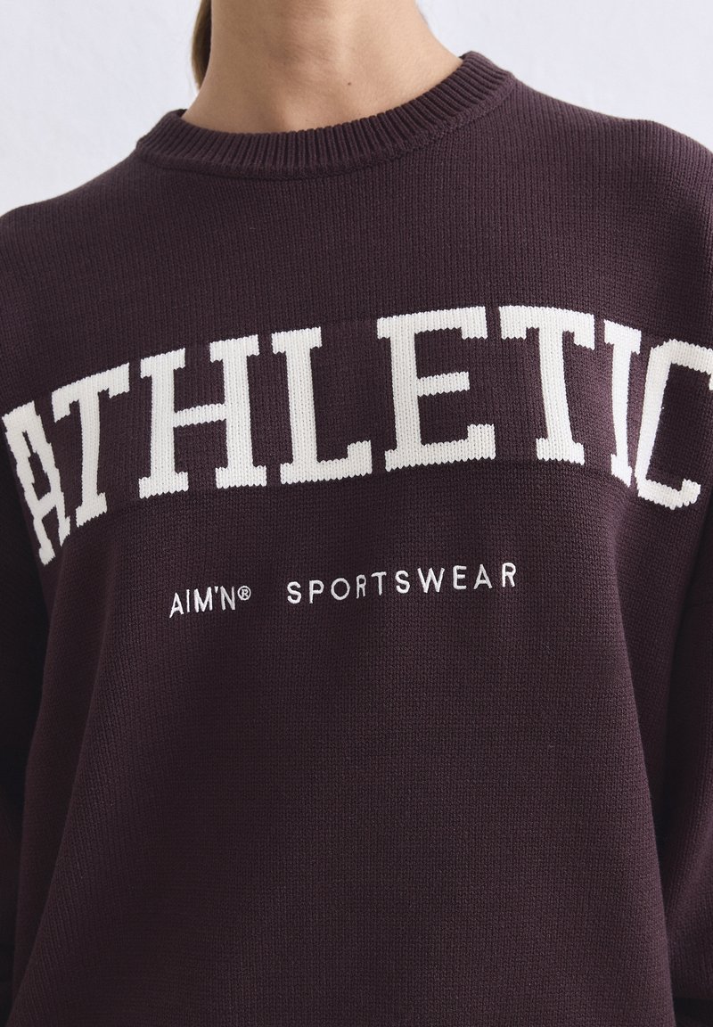 Gros plan d'un pull bordeaux avec un texte blanc "ATHLETIC" et un texte plus petit "AIM'N SPORTSWEAR" sur le devant.