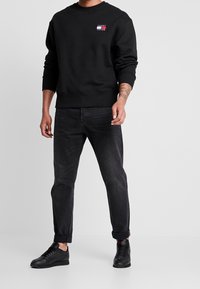 Felpa nera con polsini e orlo a costine, caratterizzata da un piccolo logo stampato in rosso, bianco e blu. Indossata con jeans scuri e sneakers nere.