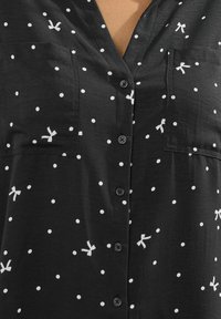 Camicia nera con bottoni decorata con pois bianchi e piccoli motivi a fiocco bianchi. Tessuto morbido con colletto classico e due taschini sul petto.