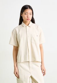 Camicia a maniche corte di colore crema con righe verticali rosse, chiusura con bottoni e taschino sul petto con un piccolo logo.