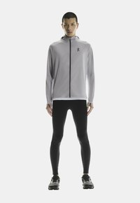 Veste de sport gris clair avec un col haut et une fermeture éclair intégrale, associée à des leggings noirs et des chaussures de course, présentant un design élégant.
