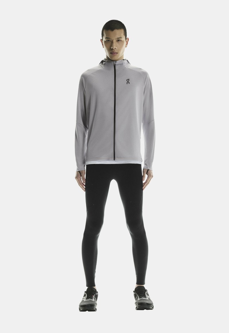 Veste de sport gris clair avec un col haut et une fermeture éclair intégrale, associée à des leggings noirs et des chaussures de course, présentant un design élégant.