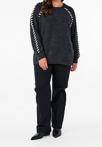 Fetter dunkelgrauer Pullover mit weißen Schnürdetails an den Ärmeln, ausgestattet mit einem gerippten Halsausschnitt und Bündchen, kombiniert mit schwarzen Hosen und Stiefeletten.