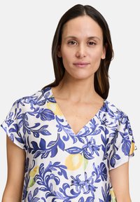 Bluse mit Blumenmuster, die einen weißen Grundton, blaue und gelbe botanische Designs, kurze Ärmel und einen V-Ausschnitt in leichtem Stoff aufweist.