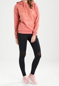 Rosa hoodie med en stor logotyp, ihopparat med svarta leggings med meshdetaljer och rosa sneakers med en texturerad yta.