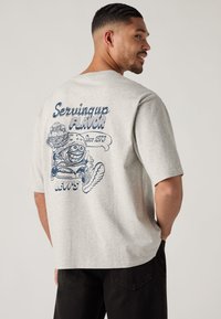 Ανοιχτό γκρι κοντομάνικο T-shirt με μπλε γραφικό στην πλάτη που απεικονίζει ένα μπεργκερ και παγωτό, με την επωνυμία "Levi's" στο κάτω μέρος.