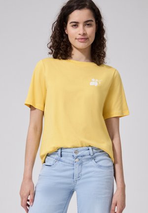 Kvinde med krøllet mørkt hår iført en gul kortærmet skjorte og lyseblå højtaljede jeans mod en ensfarvet baggrund.