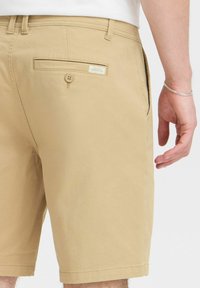 Shorts en coton beige au toucher lisse, dotés d'une poche arrière avec une fermeture à bouton et d'une petite étiquette au-dessus.