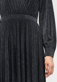 Μακρυμάνικο μαύρο φόρεμα με λαμπερή υφή, διαθέτει εφαρμοστή μέση και φαρδιά φούστα. Λεπτομέρειες με πιέτες και ελαστικά μανσέτα.