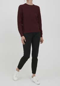 Burgunderfarbener Pullover mit strukturiertem Bommeldesign, rundem Halsausschnitt und gerippten Bündchen, kombiniert mit schwarzen Slim-Fit-Hosen und weißen Sneakern.