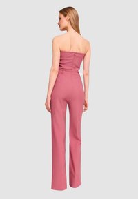 Tuta senza spalline in tessuto rosa tenue, corpetto aderente, vita con cintura e pantaloni a gamba larga. Sul retro presenta una cerniera nascosta.