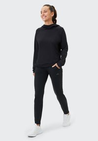 Schwarzes langärmliges Sportoberteil mit Kapuze und Taschen, kombiniert mit schwarzen engen Hosen. Abgerundet mit weißen Sportschuhen.