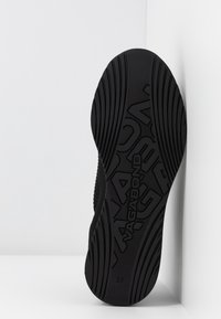 Semelle de chaussure noire avec des rainures texturées et le logo « VAGABOND » embossé au centre, positionnée à la verticale contre un mur d'angle blanc.