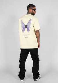 Yellow T-shirt featuring a large purple butterfly graphic on the back. Black cargo pants with pockets and white sneakers complete the outfit.