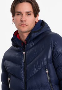 Marinblå puffjacka med chevron-quilting, dragkedjefickor och huva. Har glänsande yta och kontrasterande röd foder.