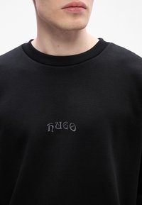 Gros plan d'une personne portant un sweat-shirt noir avec un col côtelé et "hugo" brodé en petites lettres grises sur la poitrine.