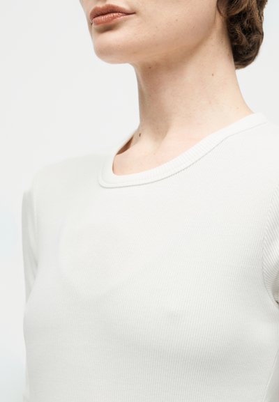 Γυναίκα με λευκό ριμπ πλεκτό μακρυμάνικο πουκάμισο, που φαίνεται το λαιμό και μέρος του προσώπου με φυσικό μακιγιάζ και καστανά μαλλιά που είναι τραβηγμένα προς τα πίσω.