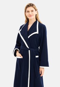 Frau trägt einen marineblauen Bademantel mit weißem Besatz, tailliertem Gürtel und Taschen, stehend vor einem schlichten, hellen Hintergrund.