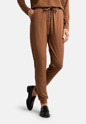 Kobieta w brązowych spodniach dresowych ze sznurkiem i czarnych mokasynach, stojąca z jedną ręką w kieszeni na białym tle.