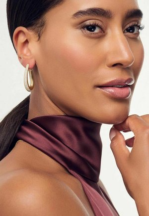 Närbild av en kvinna som bär en djupt bourgogny satintopp. Hon har gyllene hoopsmycken och en slät, glansig hy, med en subtil, naturlig sminklook.