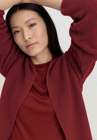 Bordeauxfarbener, strukturierter Cardigan über einem passenden T-Shirt. Der Cardigan hat gerippte Ärmel und eine lockere Passform, mit einer glatten Stoffoberfläche.
