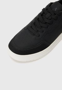Black sneaker with a smooth leather upper and suede accents. Features perforated detailing, black laces, and a white rubber sole.