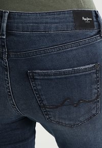 Mörkblå denimjeans med en synlig bakficka som har en svart etikett märkt "Pepe Jeans London." Tyget har en slät textur.