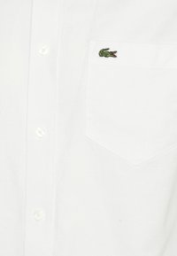 Vit skjorta med knappar och framficka broderad med en liten grön krokodil-logotyp.