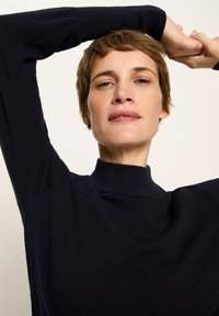 Frau mit kurzem braunem Haar, die einen schwarzen Rollkragenpullover trägt und beide Arme über den Kopf hebt, vor einem schlichten, hellen Hintergrund.