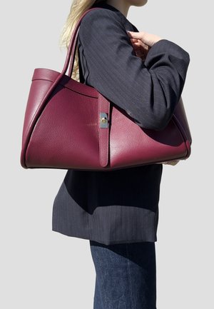 Persona con blazer scuro e jeans porta una grande borsa a tracolla in pelle color granata e lavorata su uno sfondo neutro.