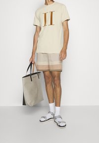 Hombre con camiseta beige con estampado II, pantalones cortos a rayas beige y marrón, calcetines blancos con sandalias plateadas, sosteniendo una bolsa de mano beige y negra.