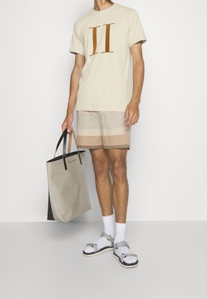 Hombre con camiseta beige con estampado II, pantalones cortos a rayas beige y marrón, calcetines blancos con sandalias plateadas, sosteniendo una bolsa de mano beige y negra.