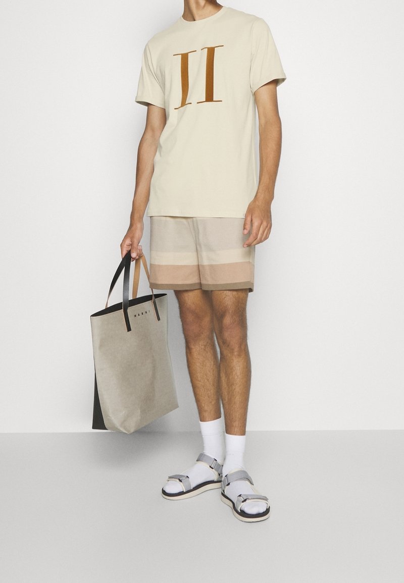 Hombre con camiseta beige con estampado II, pantalones cortos a rayas beige y marrón, calcetines blancos con sandalias plateadas, sosteniendo una bolsa de mano beige y negra.