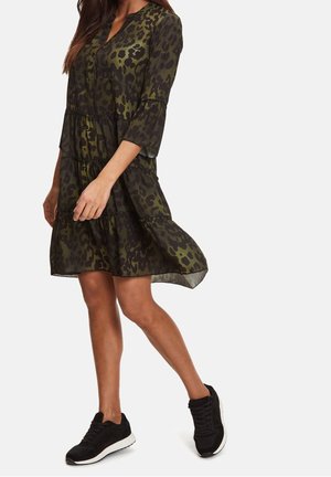 Femme marchant portant une robe mi-longue à imprimé léopard vert et noir, manches trois-quarts, et des baskets noires.