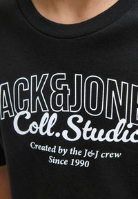 Crni pamučni majica s okruglim izrezom, s odvažnim bijelim natpisom: "BLACK&JONES Coll. Studio" i manjim tekstom ispod.