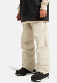 Beige waterdichte broek met een losse pasvorm, voorzien van stikseldetails en verstelbare enkelsluitingen. Gekleed met zwarte instappers.